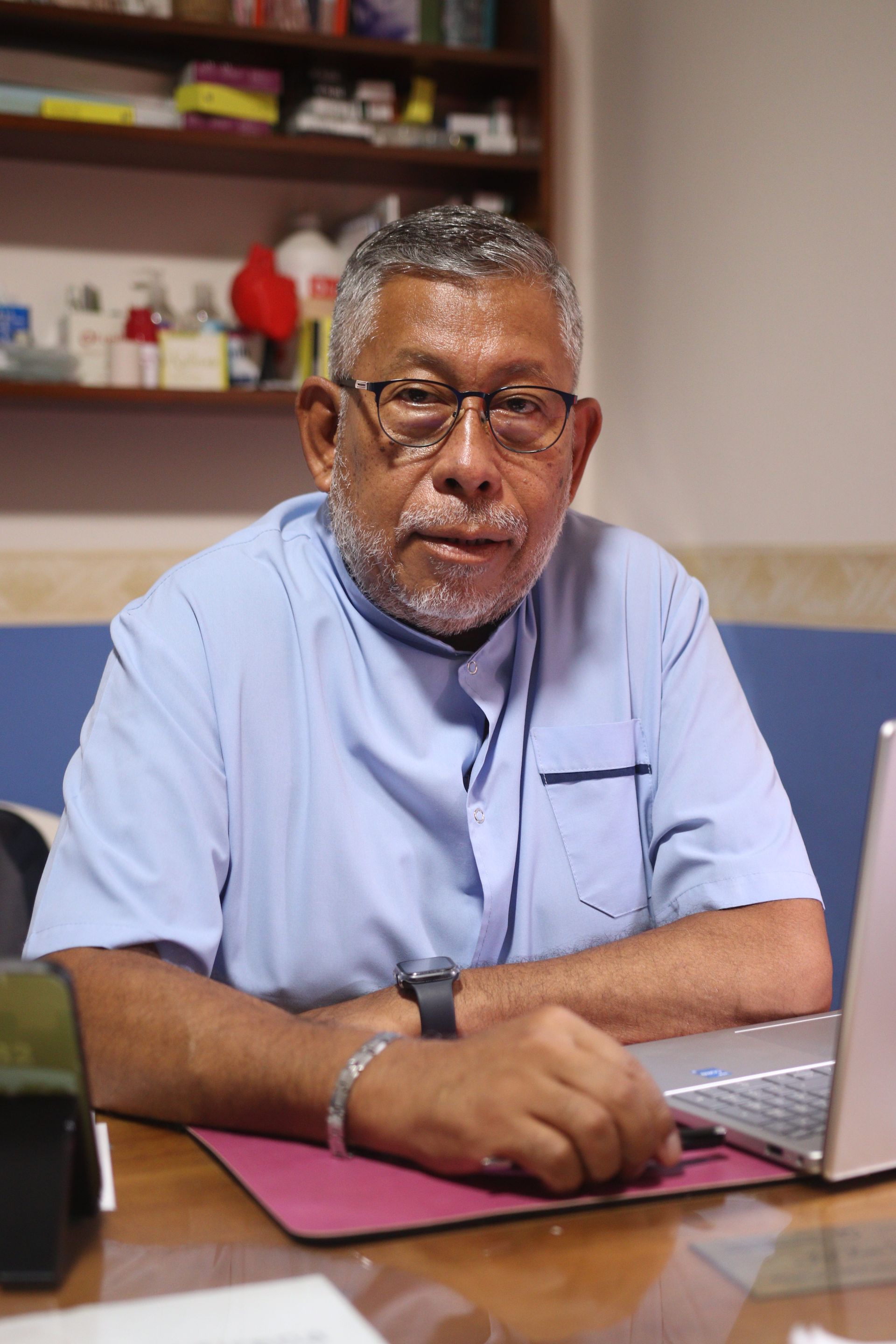Hombre sentado en un escritorio con una computadora portátil, vistiendo una camisa azul claro, en un entorno de oficina.