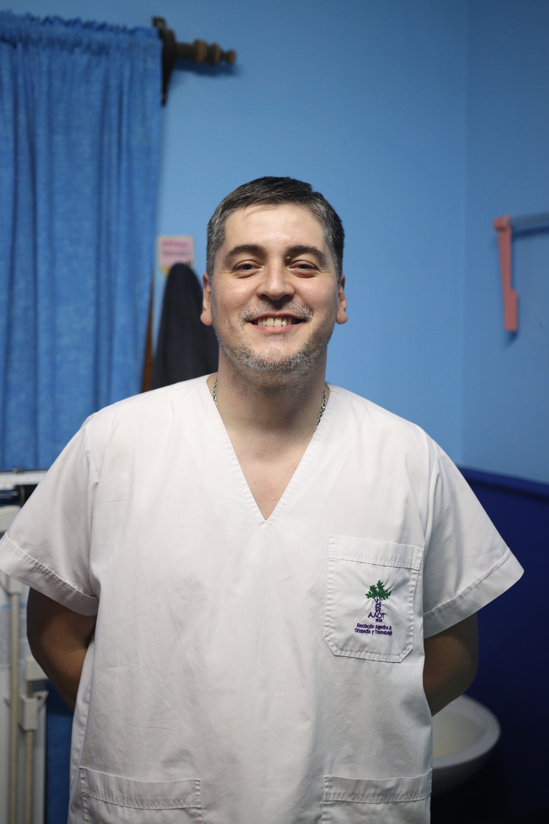 Una persona sonriente con uniforme blanco está de pie en una habitación azul con una cortina y un lavabo detrás.
