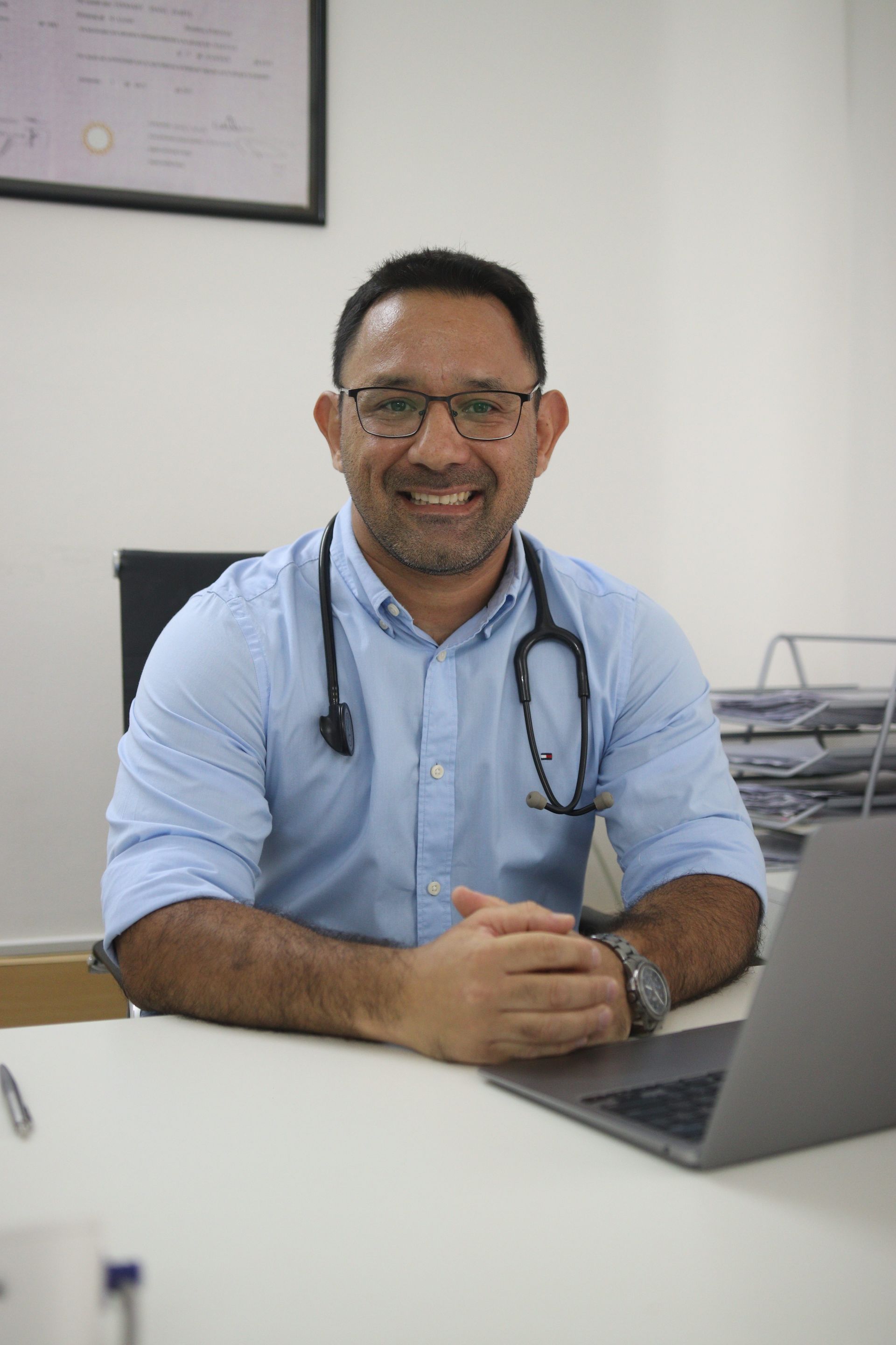 Un médico sonriente, con camisa azul claro y estetoscopio, está sentado en un escritorio