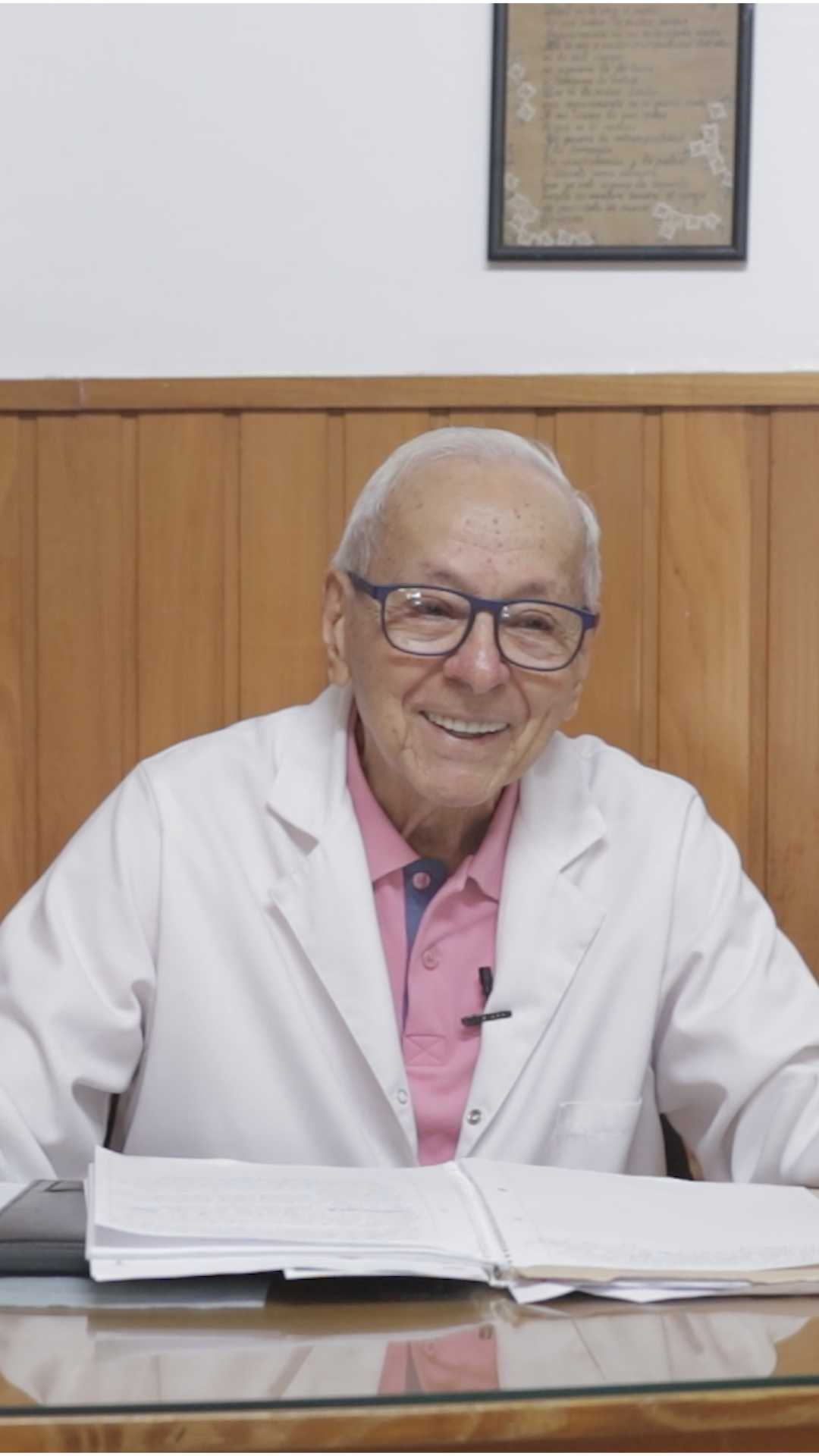Un médico sonriente, con bata blanca y camisa rosa, sentado detrás de un libro abierto en un escritorio de una oficina.