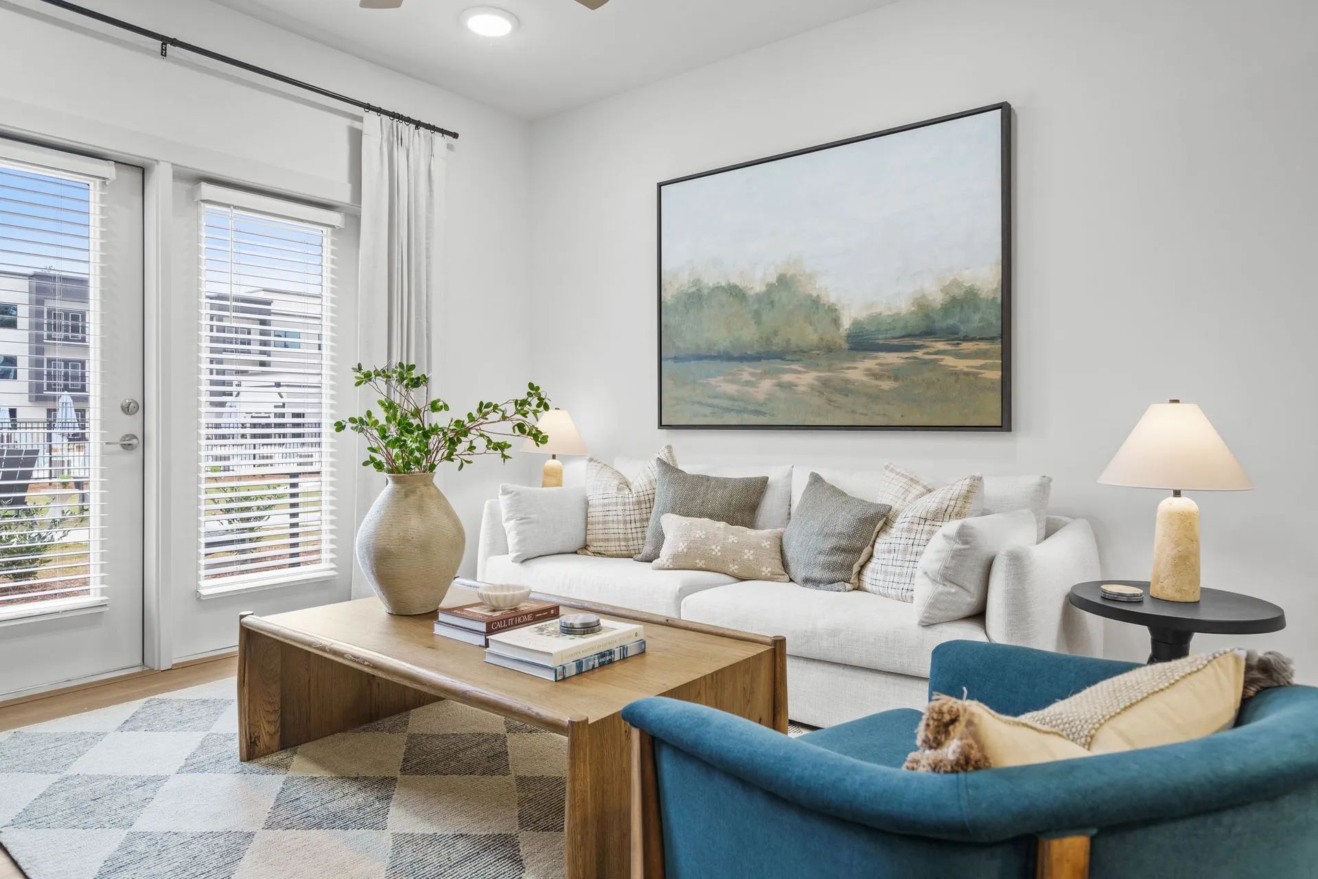 Living room with white sofa, blue armchair, and art. Large windows and wooden coffee table.