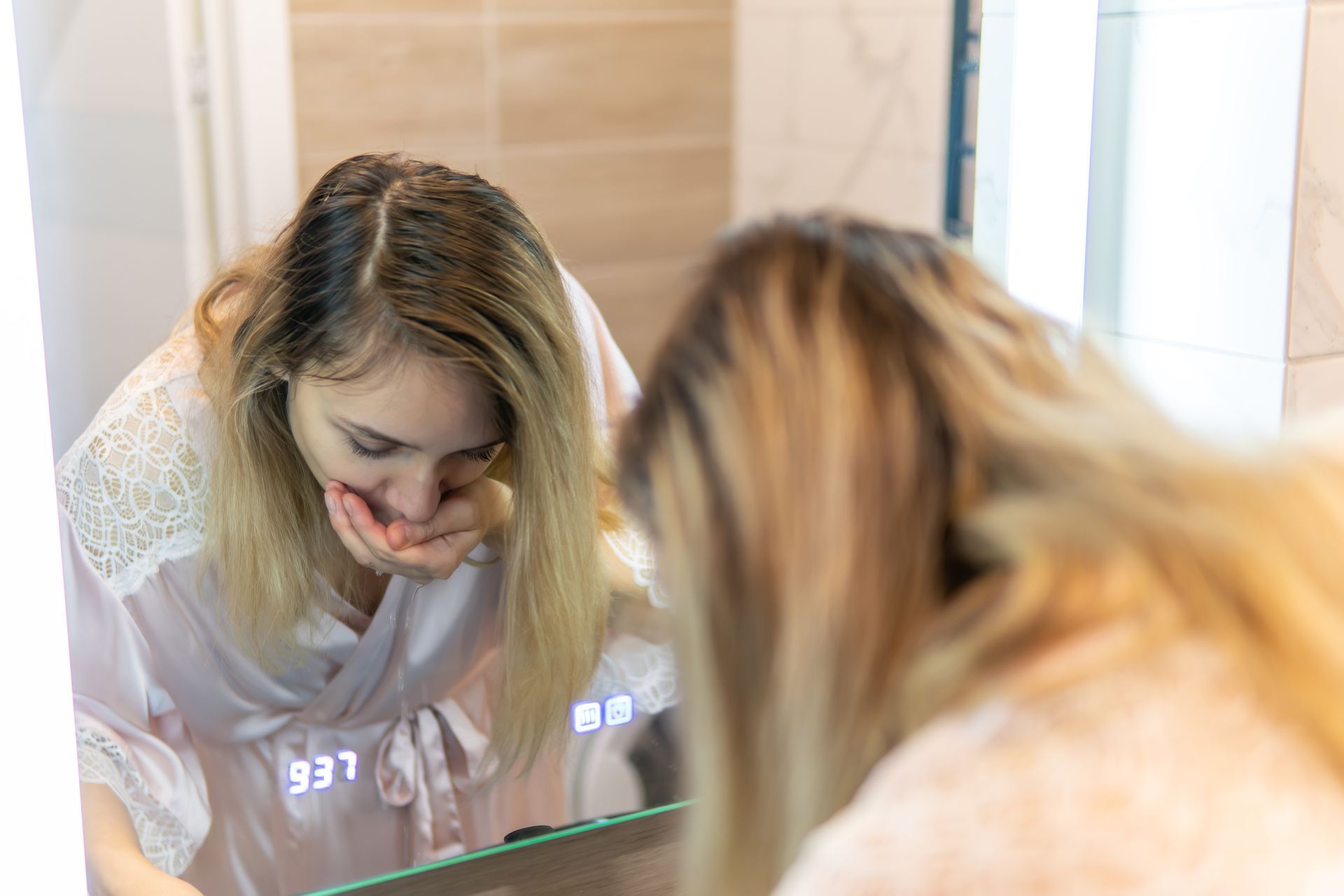 Person leaning over a sink, looking at their reflection in a bathroom mirror