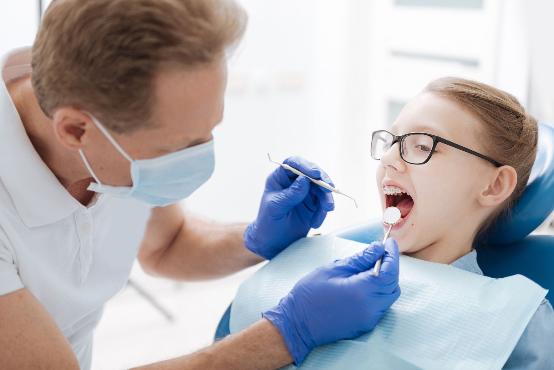 Dentist examining a child's open mouth with a dental tool, wearing a mask and gloves