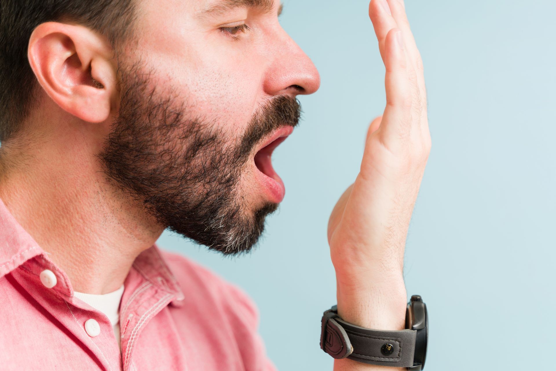 Man with beard and pink shirt yawns beside his raised hand against a light blue background