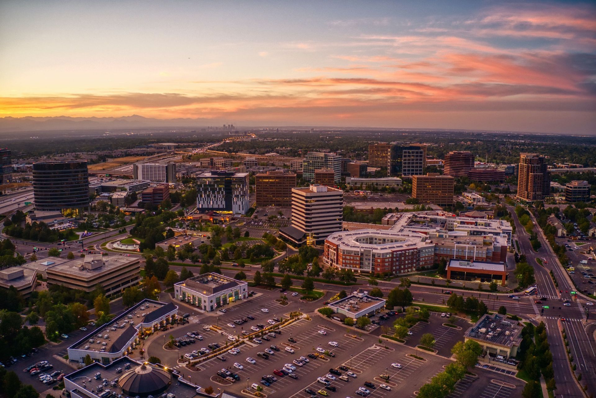 Aerial view of a city at sunset with colorful sky; includes buildings, parking lots, and trees.