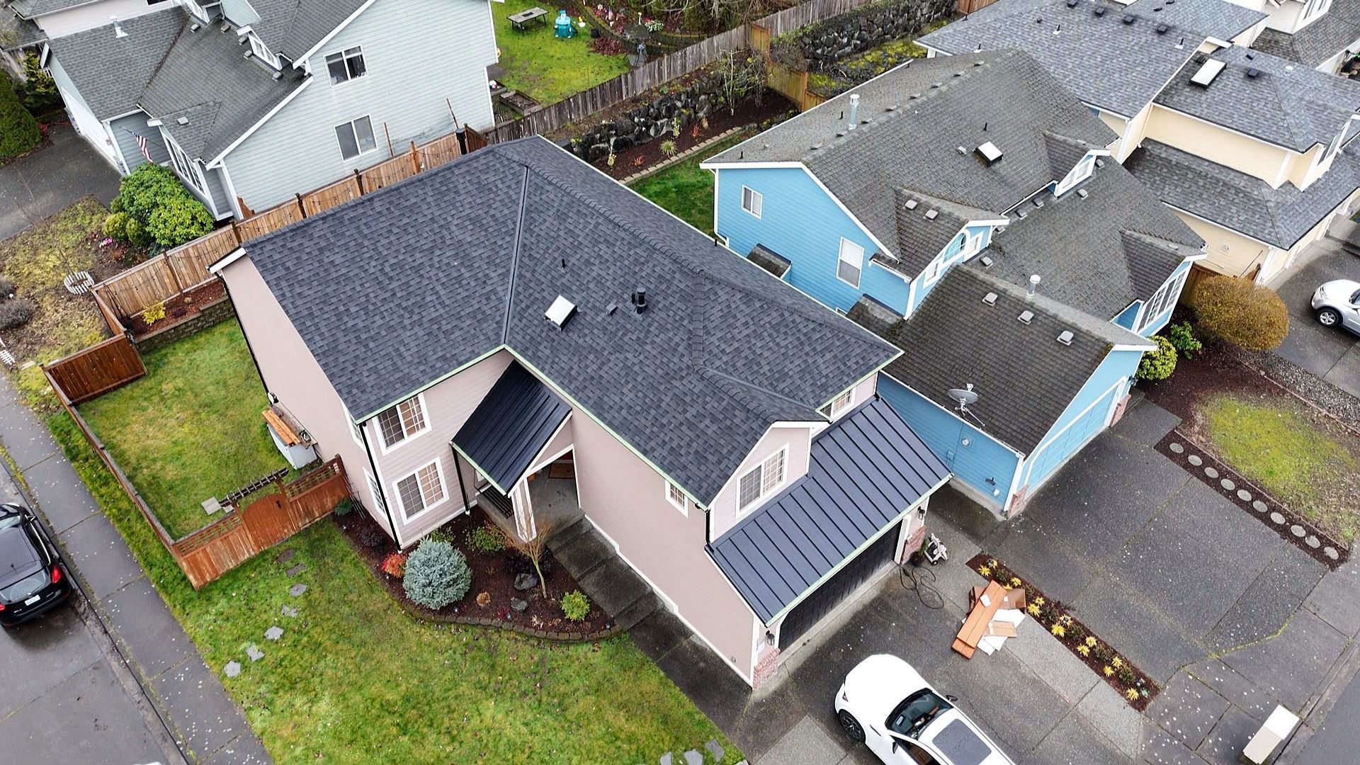 Aerial view of a suburban house with a dark roof, beige walls, and a driveway beside neighboring homes.