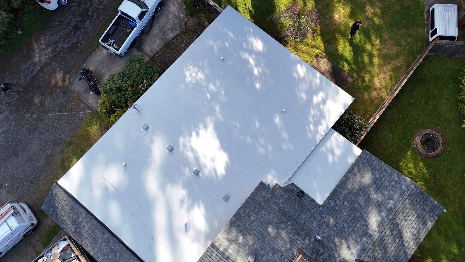 Aerial view of a white-roofed house with parked cars and trees around it.