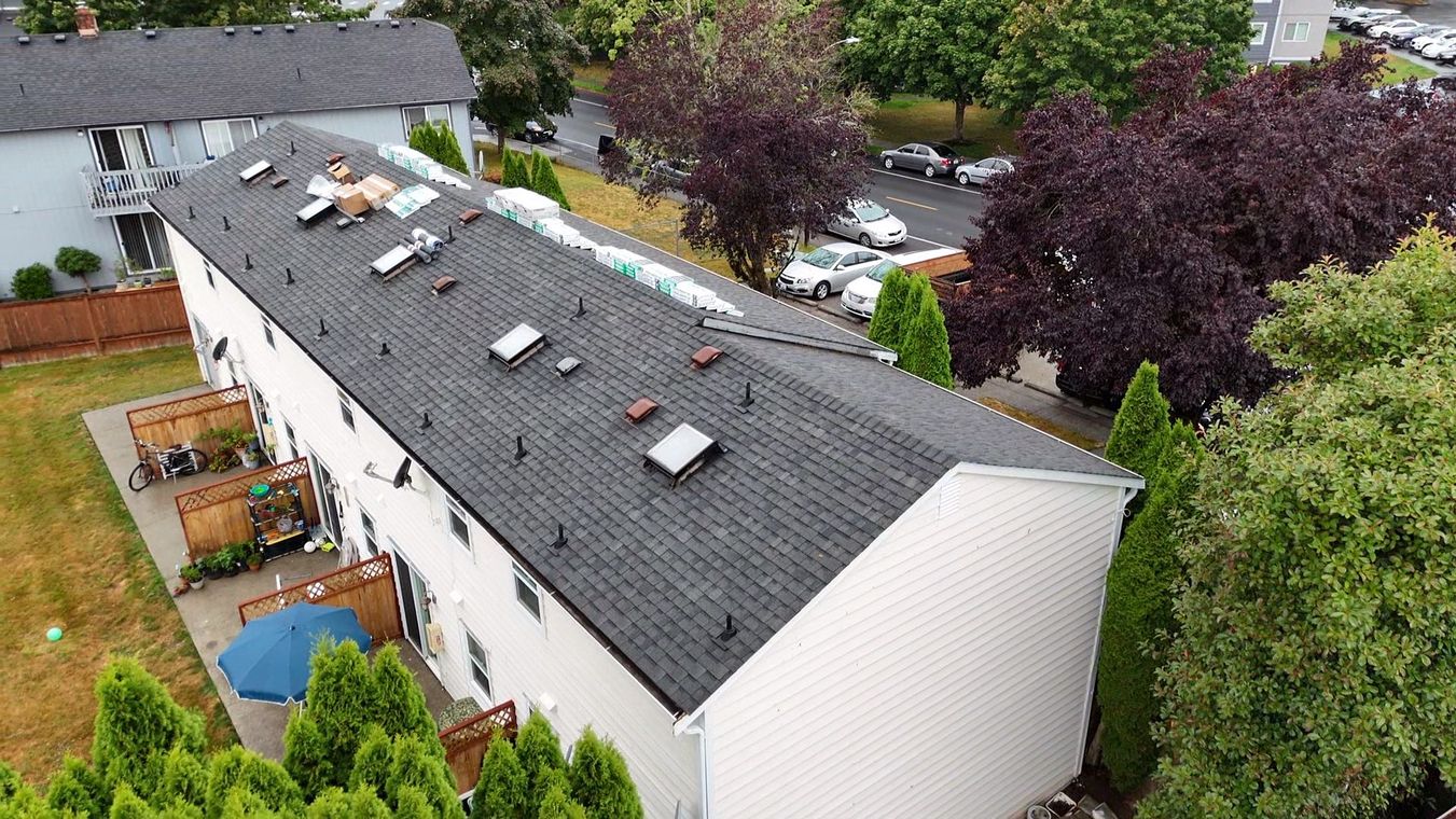 Aerial view of a long white house with a dark roof, skylights, and surrounding trees and gardens.
