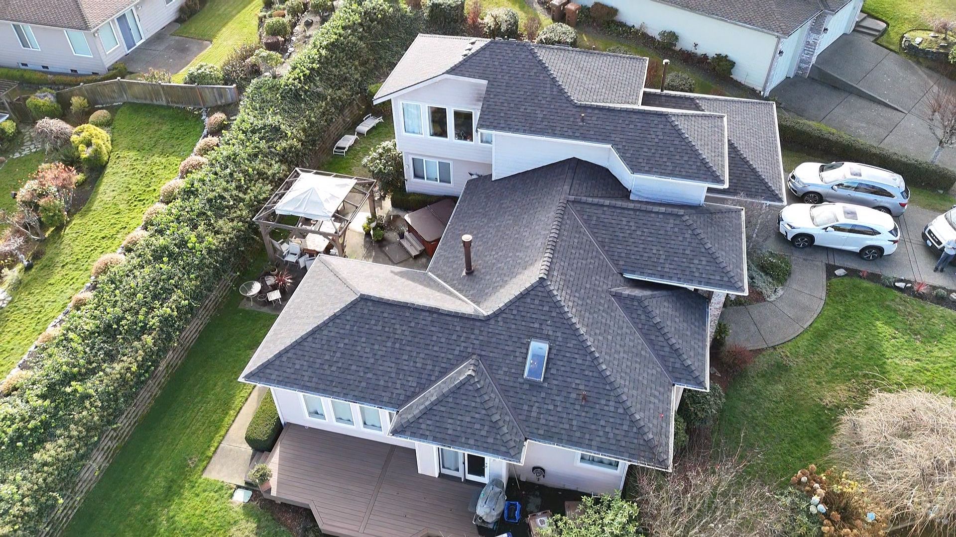 Aerial view of a suburban house with a dark roof, driveway, and surrounding green lawn and hedges