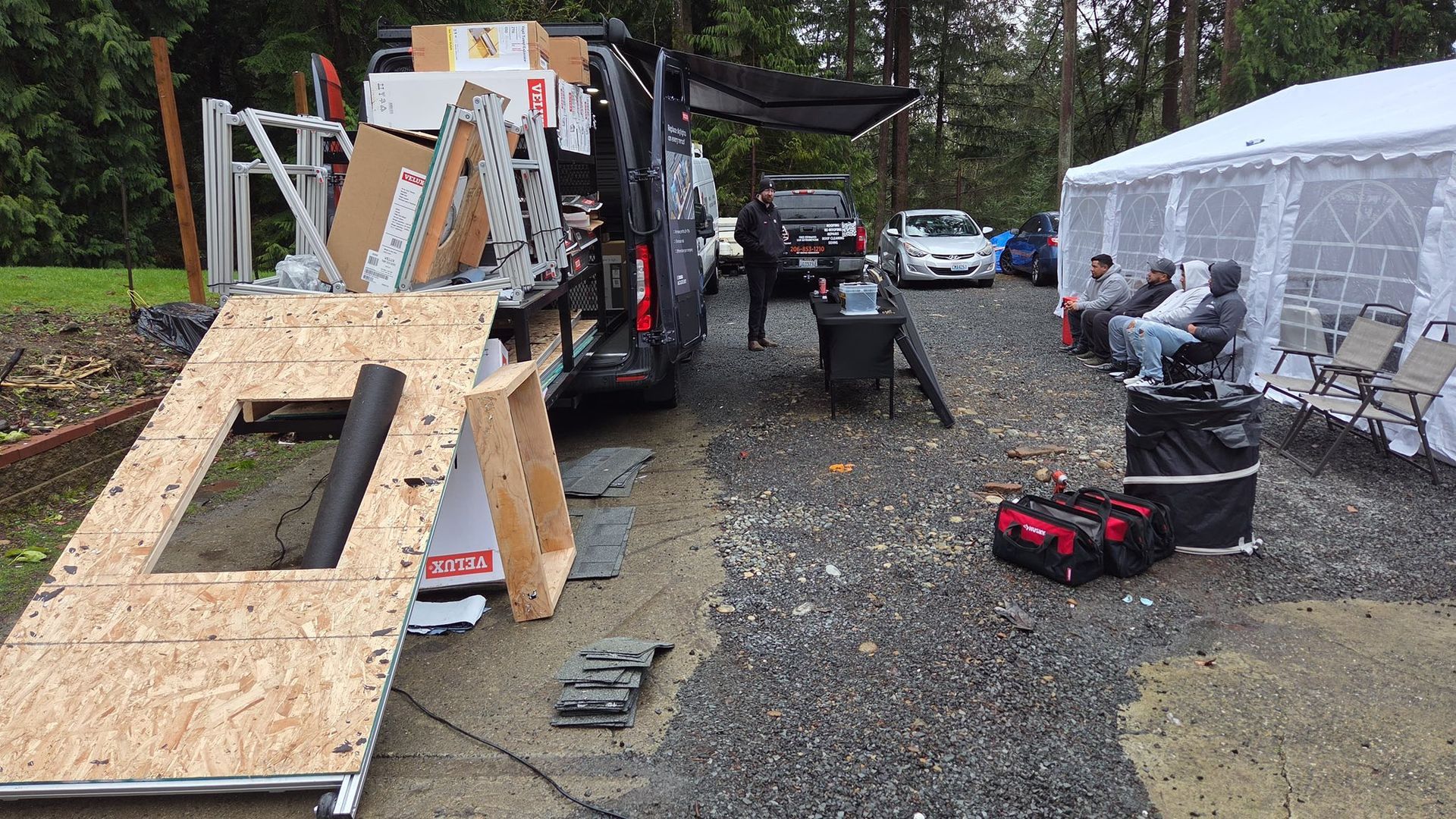 People moving equipment on a gravel driveway beside a truck and white tents after rain
