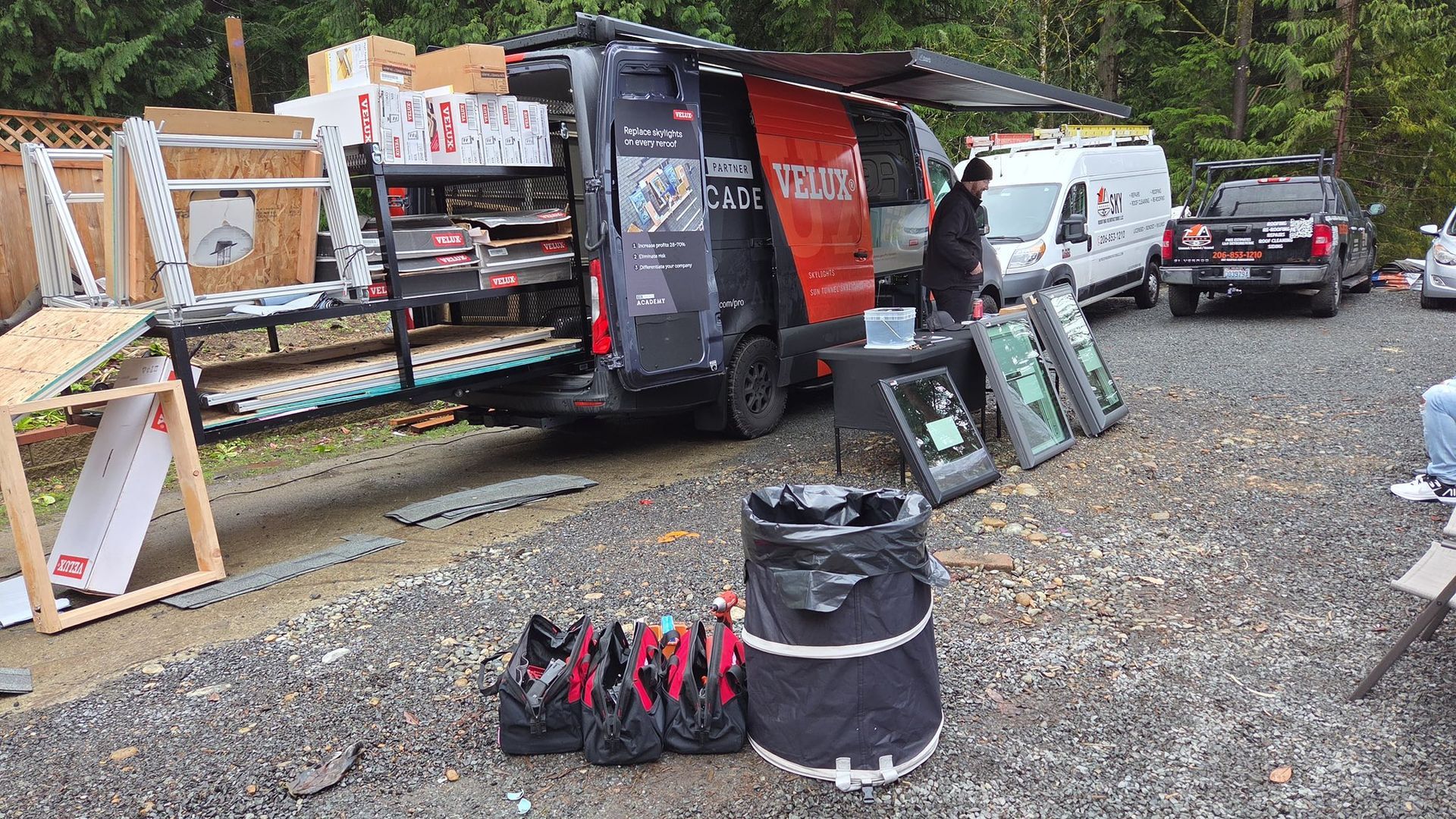 Outdoor market stall with black van, framed signs, white van, and backpack display on gravel lot