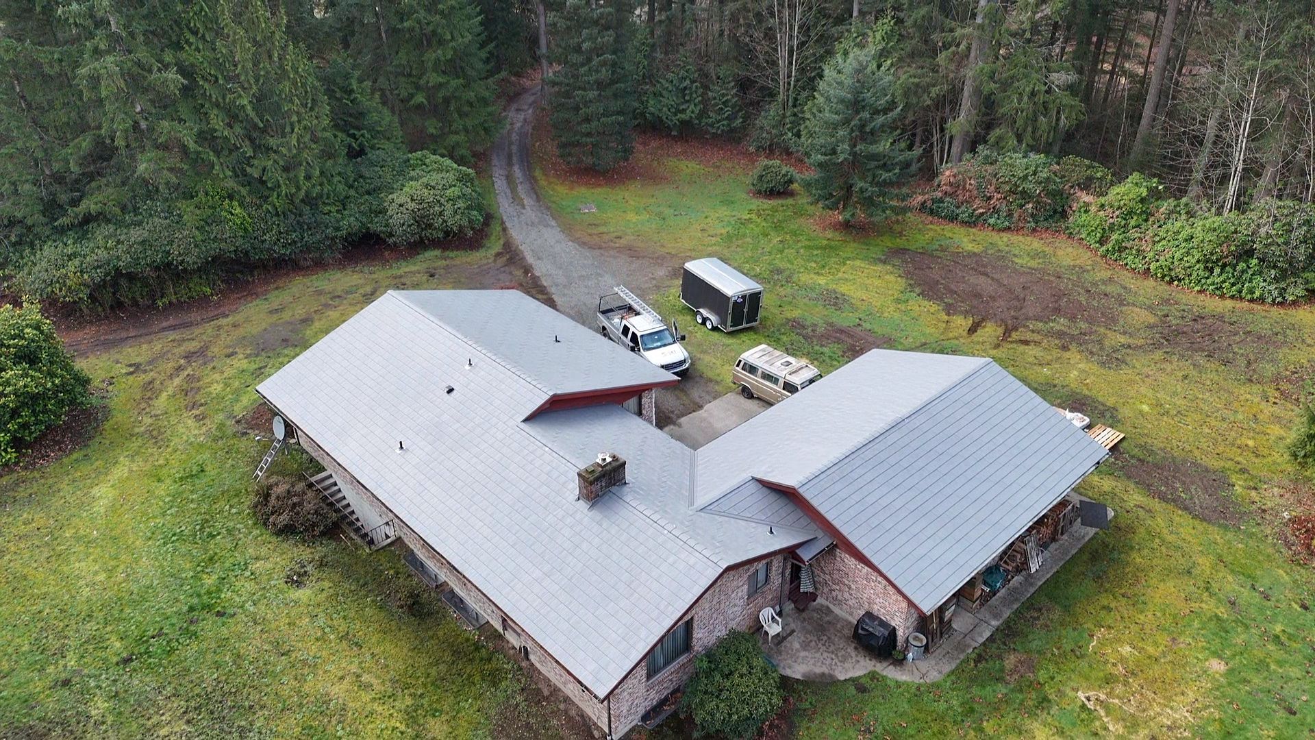 Aerial view of a rural house with gray roofs, a driveway, and surrounding trees and grass.