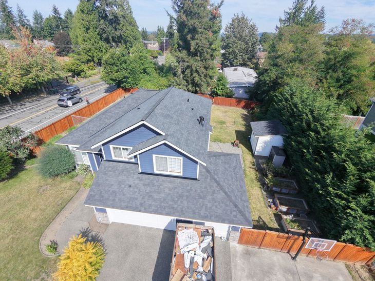 Aerial view of a house with a gray shingle roof, driveway, and surrounding trees and neighboring homes