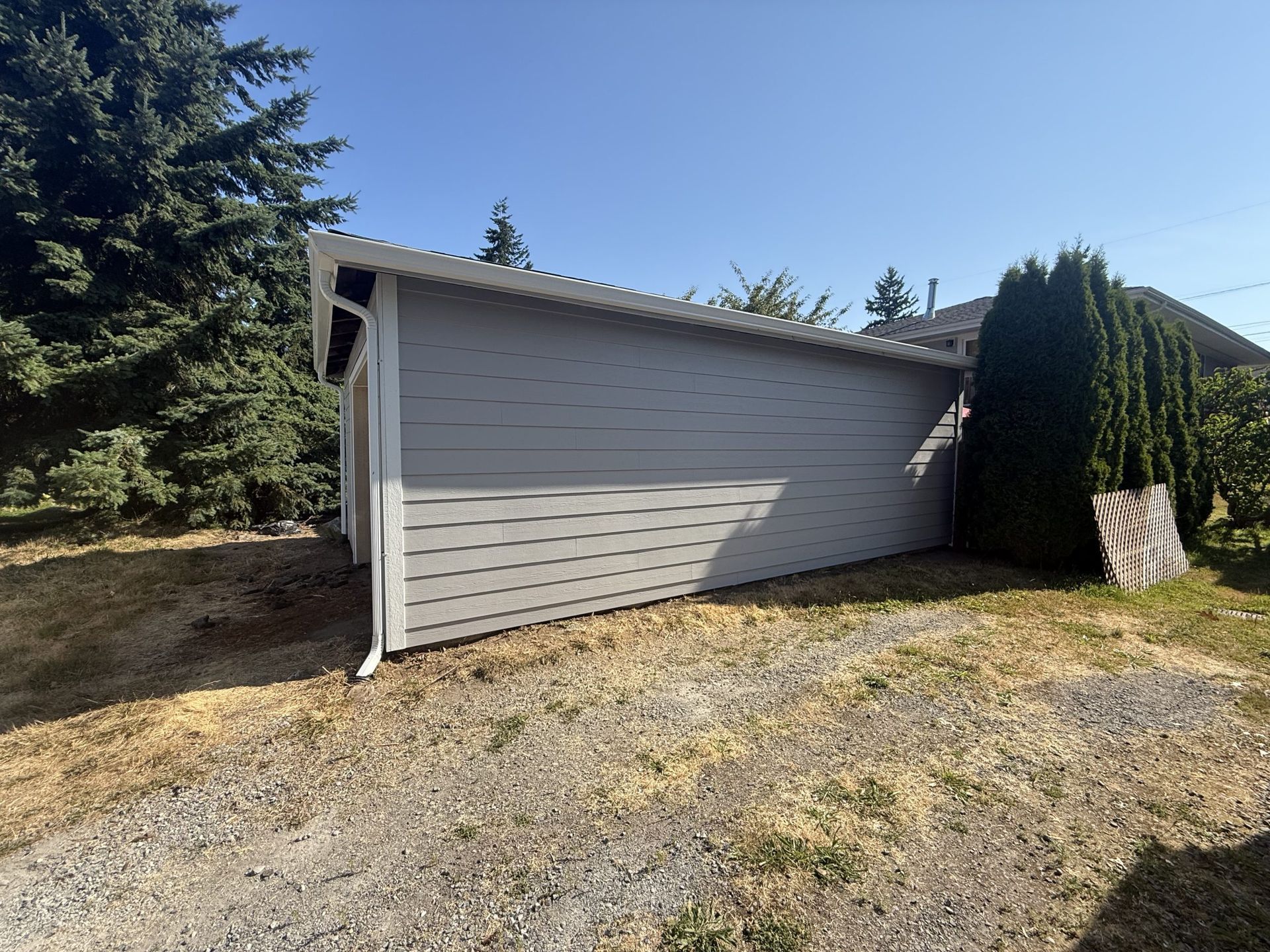 Gray detached garage beside evergreen trees on a gravel driveway under a clear blue sky