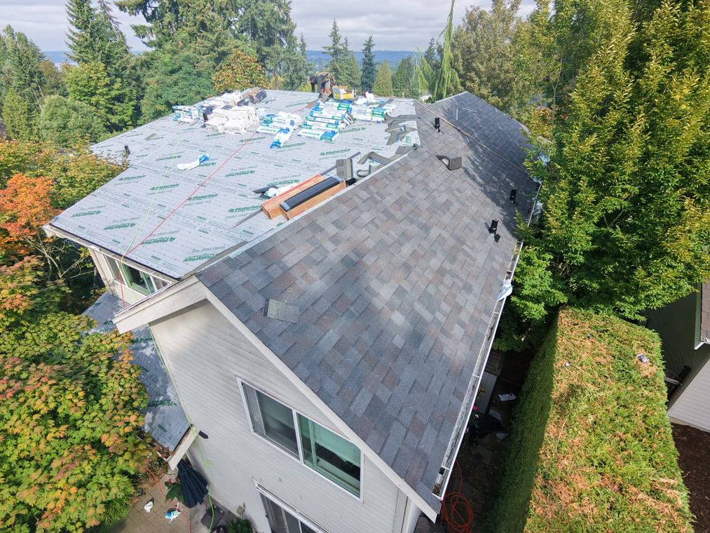 Aerial view of a house with gray roof, wooden deck, and surrounding trees near a street with parked cars