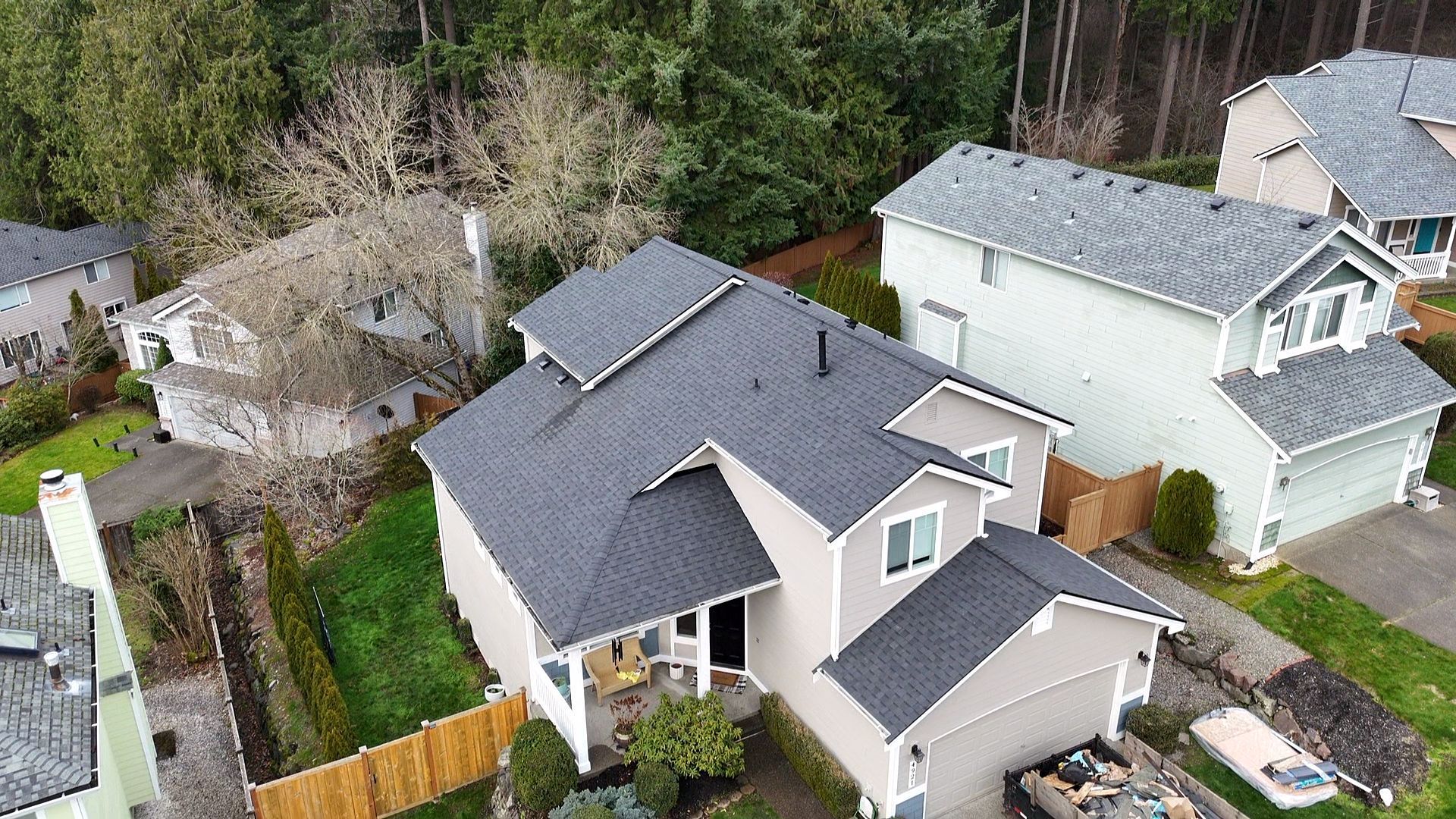 Aerial view of suburban houses with gray roofs, green yards, and a wooded backdrop