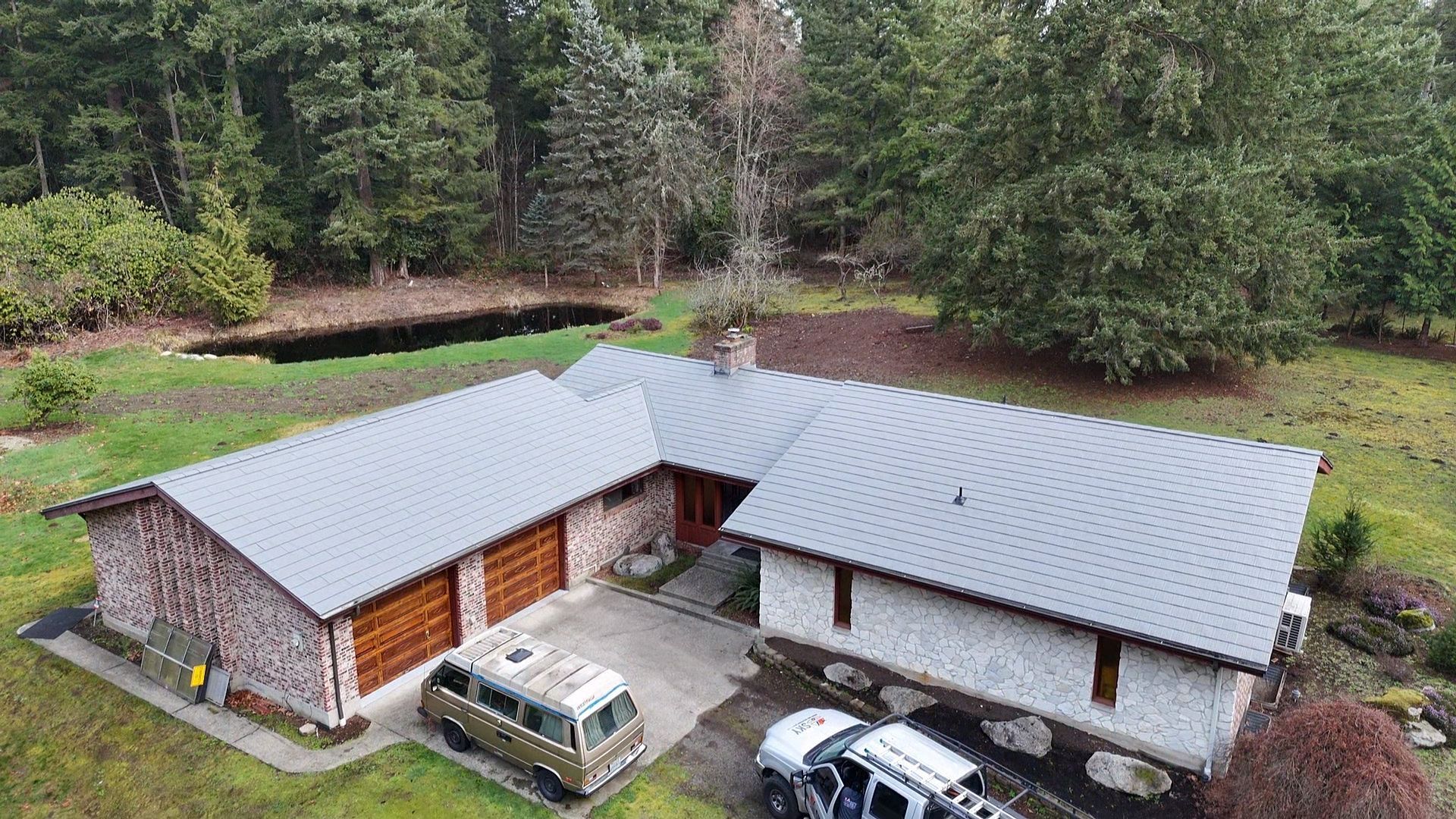 Aerial view of a modern house with gray roofs, stone walls, and parked vehicles beside a wooded yard.