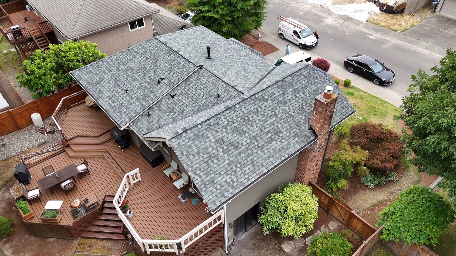 Aerial view of a gray-roofed house with a wooden deck, brick patio, and parked cars nearby.