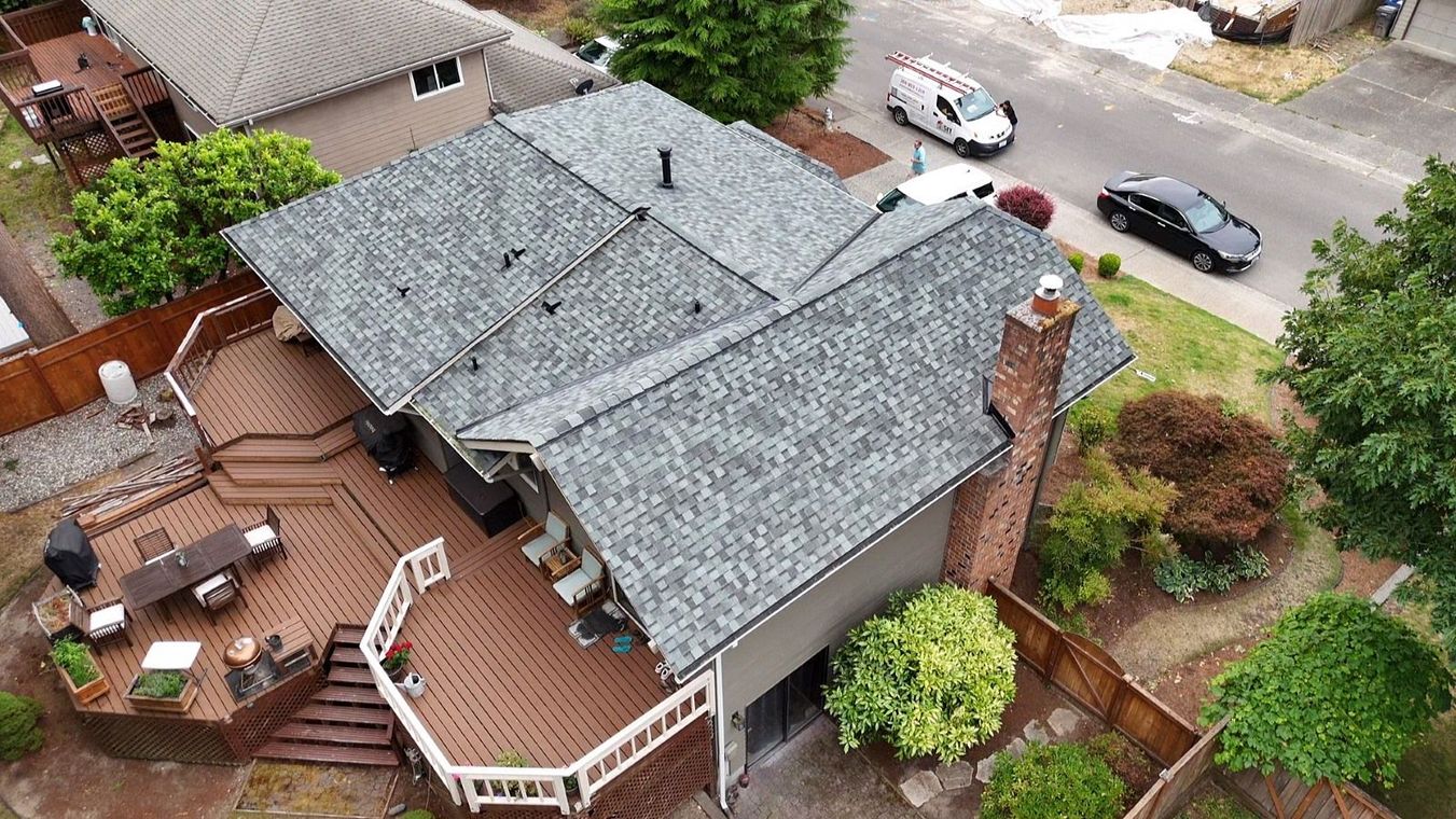 Aerial view of a house with gray roof, wooden deck, and surrounding trees near a street with parked cars
