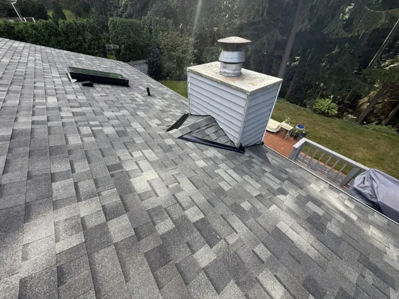 Gray shingle roof with a white vented chimney, small roof opening, and surrounding trees on a sloped house roof