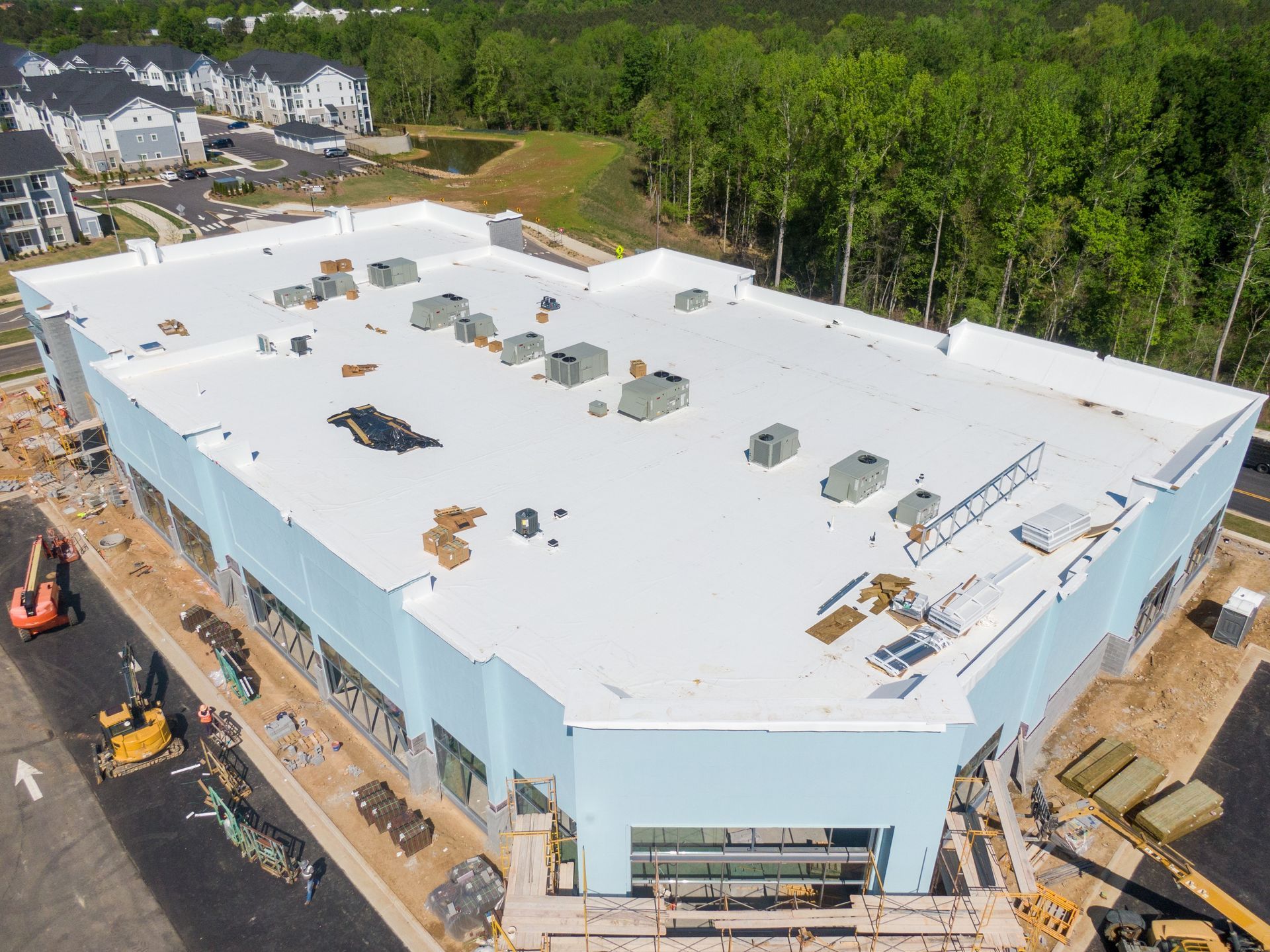 Aerial view of a large white-roofed building under construction beside a wooded area and parking lot