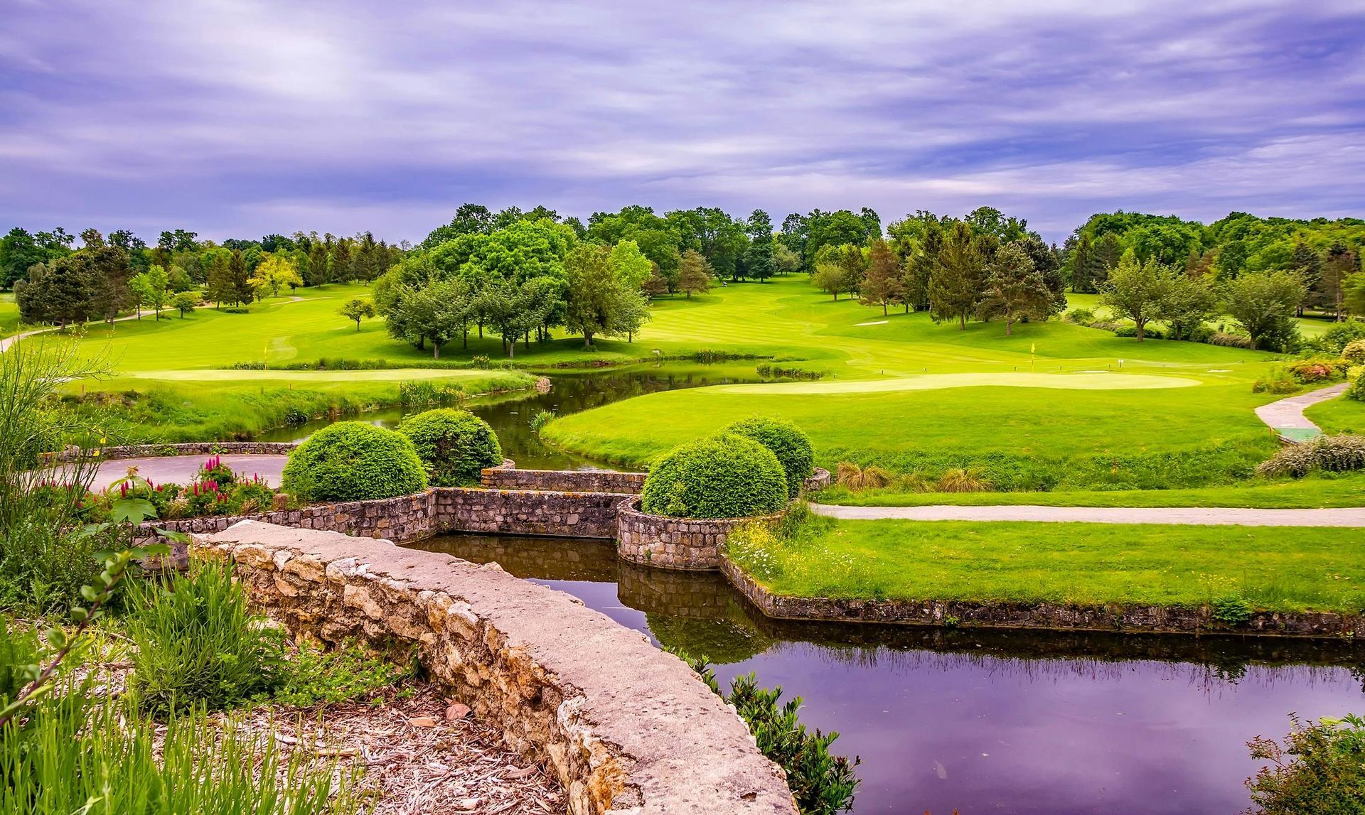 Lush green golf course with a stone bridge, winding path, and pond under a purple sky