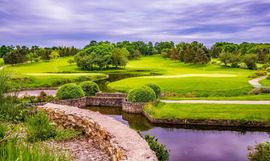 Lush green golf course with a stone bridge, winding path, and pond under a purple sky