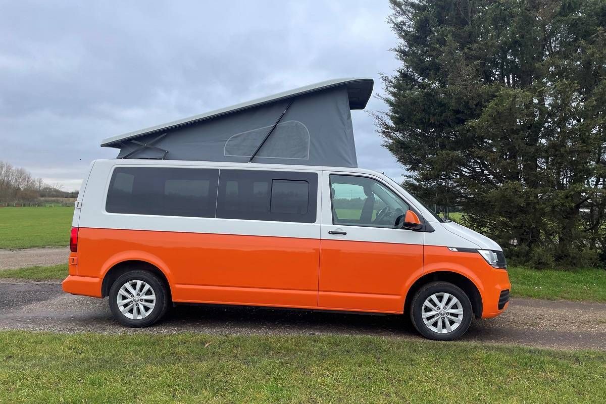 An orange and white van with a pop up roof is parked in a grassy field.