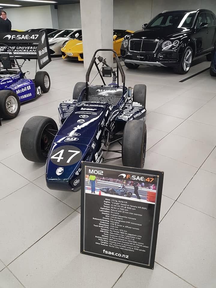 Formula race car on display in a museum, with a black information sign and other vehicles in the background