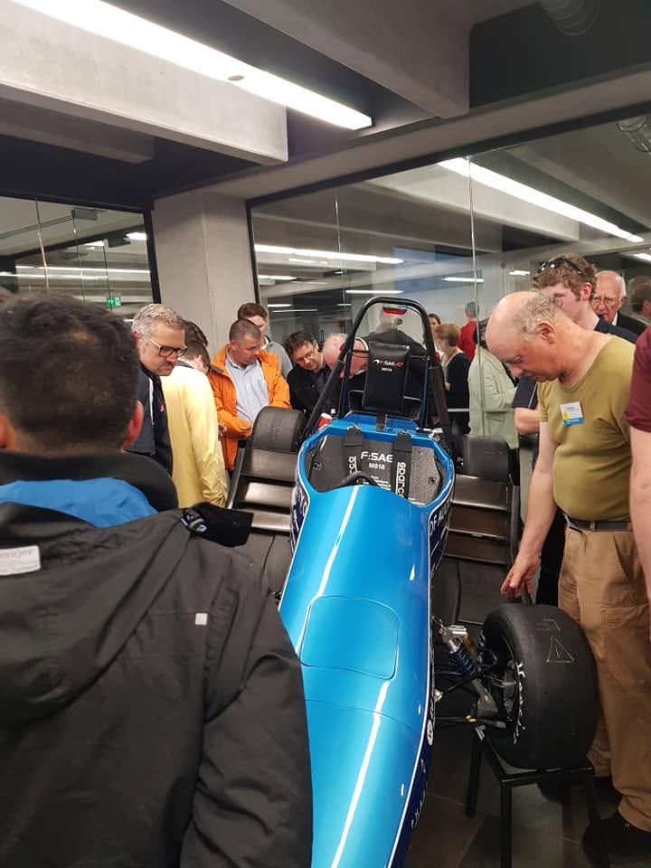 Blue race car surrounded by people in a garage, with several men inspecting it closely.