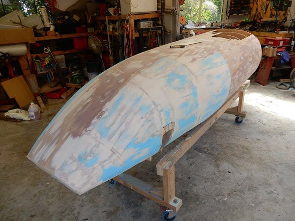 Partially sanded boat hull on a stand in a workshop, with blue and brown primer patches