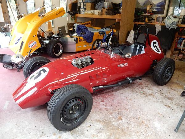 Red vintage race car in a garage, with a yellow race car behind it.