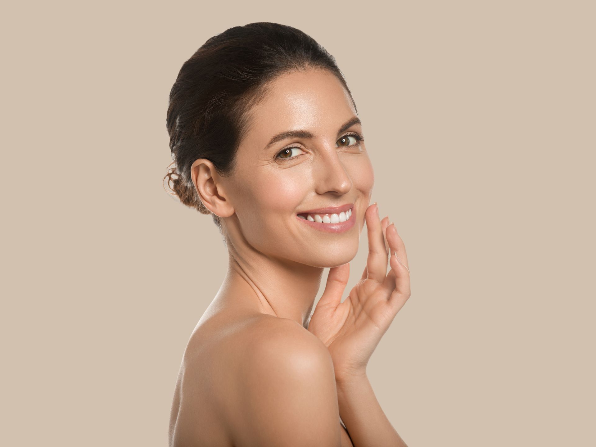 Woman with radiant skin smiles, touching her cheek, brown hair pulled back, against a beige background.
