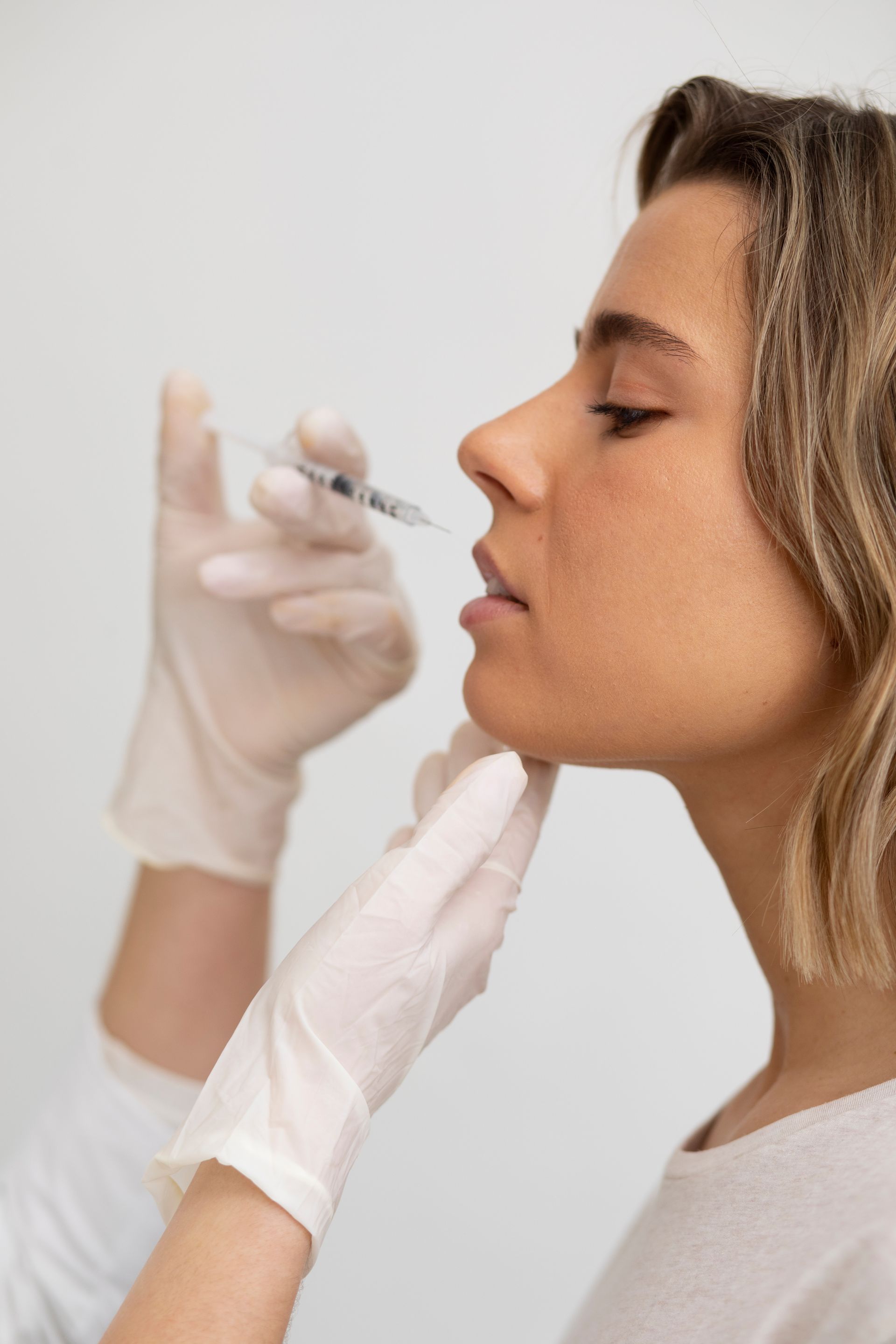 Woman receiving lip injection; doctor's gloved hands hold syringe near lips. Light-skinned, neutral background.