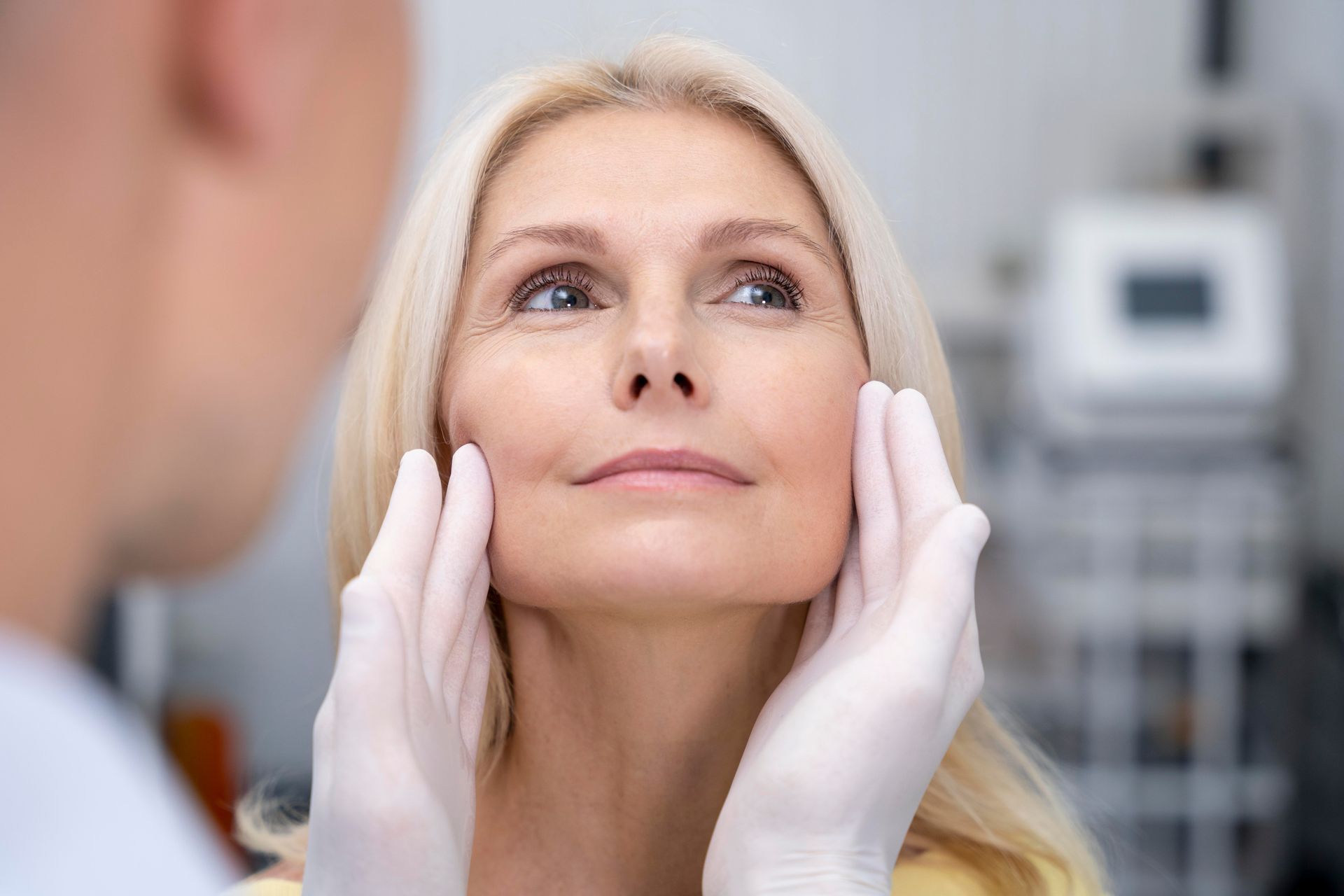 Woman's face being examined by gloved hands, possibly a doctor's, in a clinic.