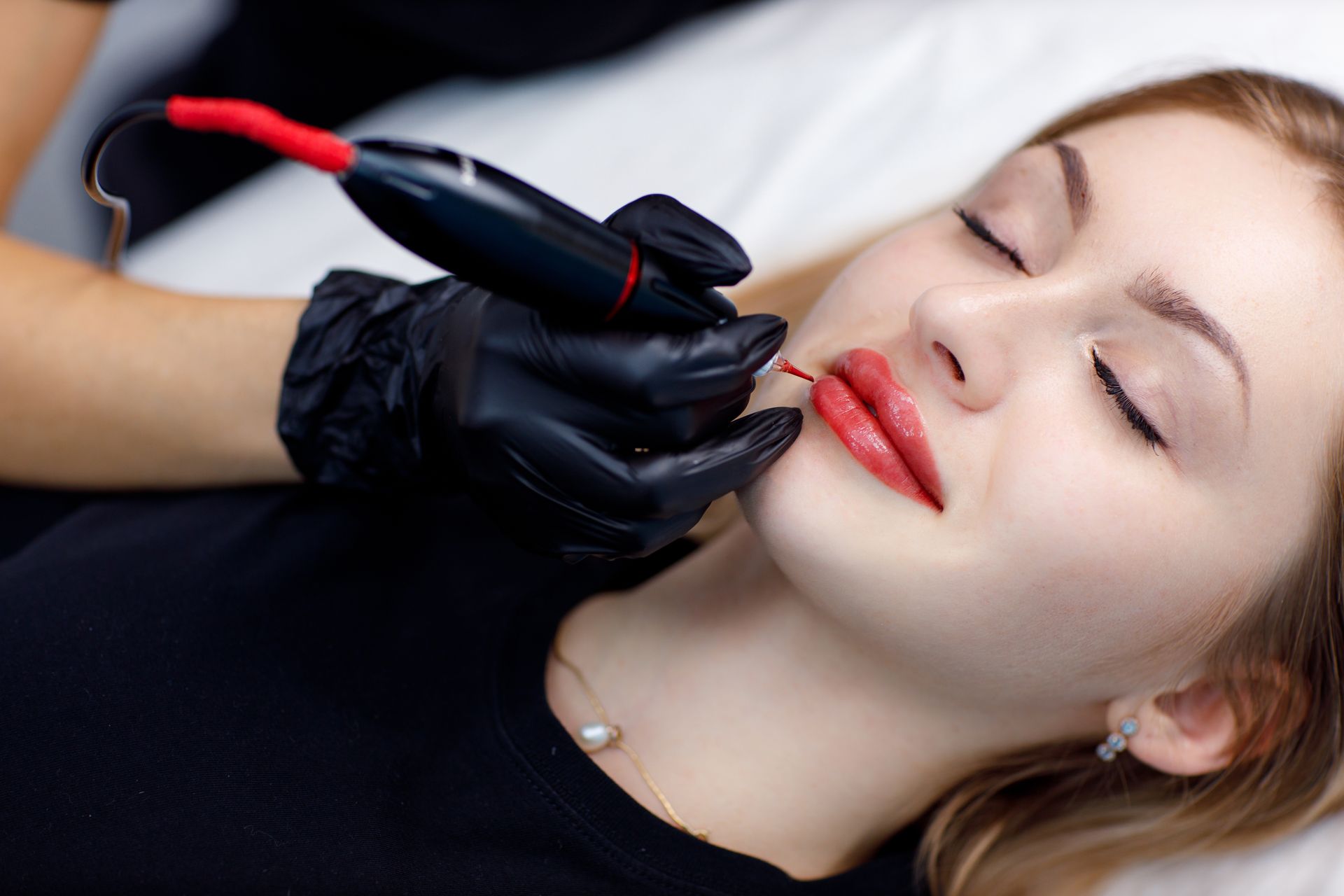 Woman getting lip tattoo, red ink applied with machine. Black-gloved hand, closed eyes, medical setting.