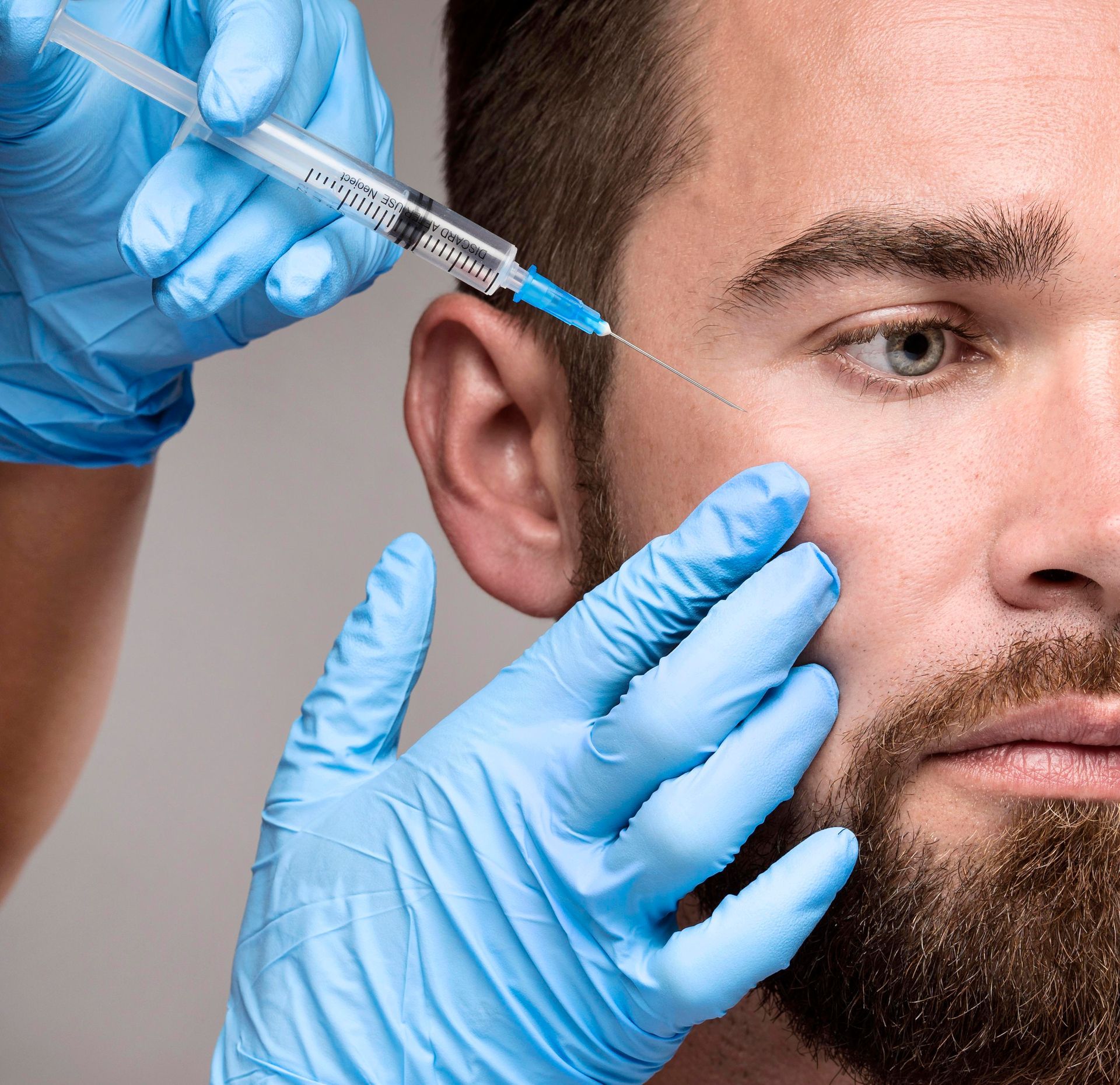 Man receiving cosmetic injections in the face; blue-gloved hands hold a syringe near his eye.