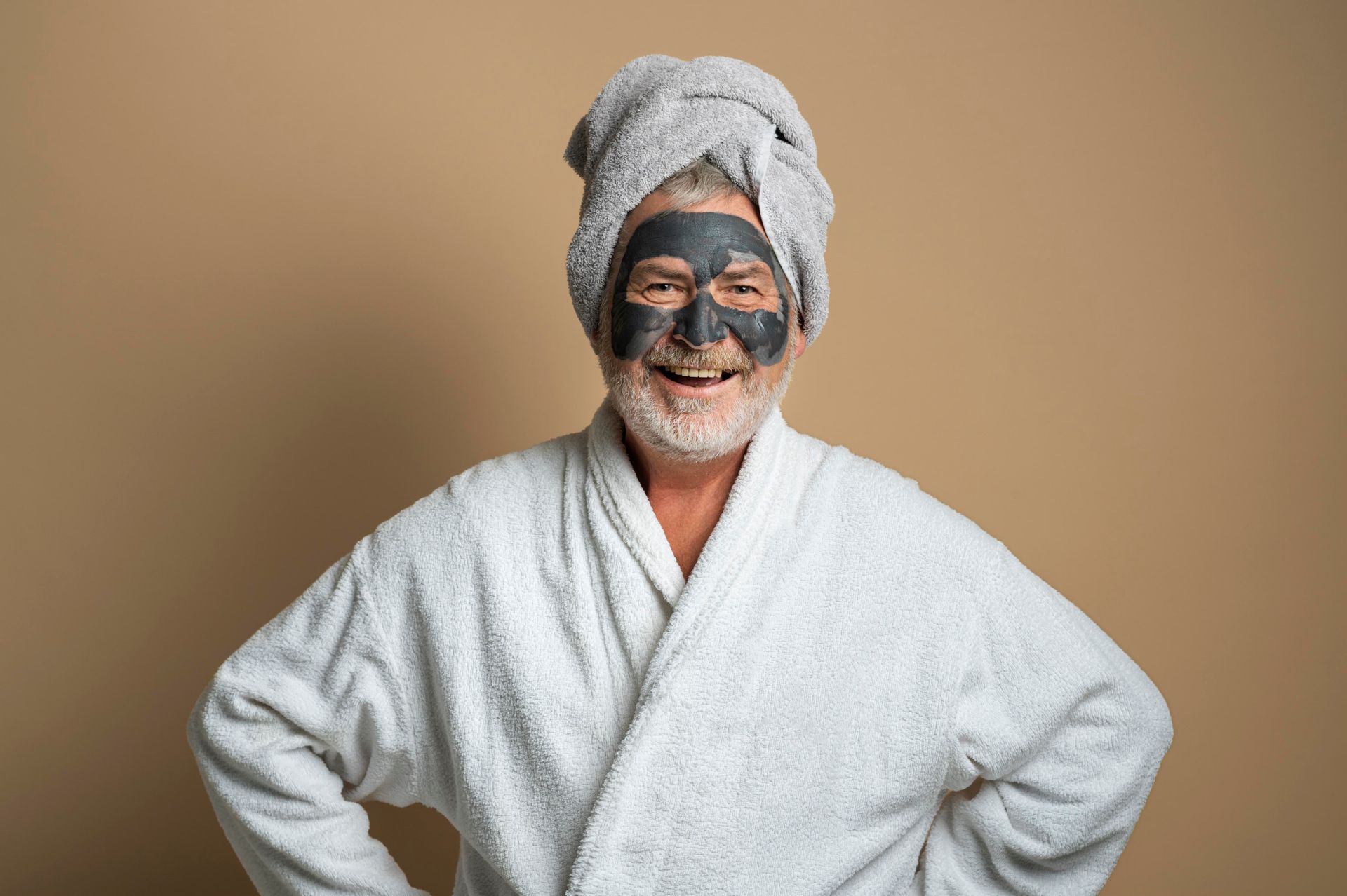 Person in a white bathrobe and towel turban with a facial mask, smiling against a beige background