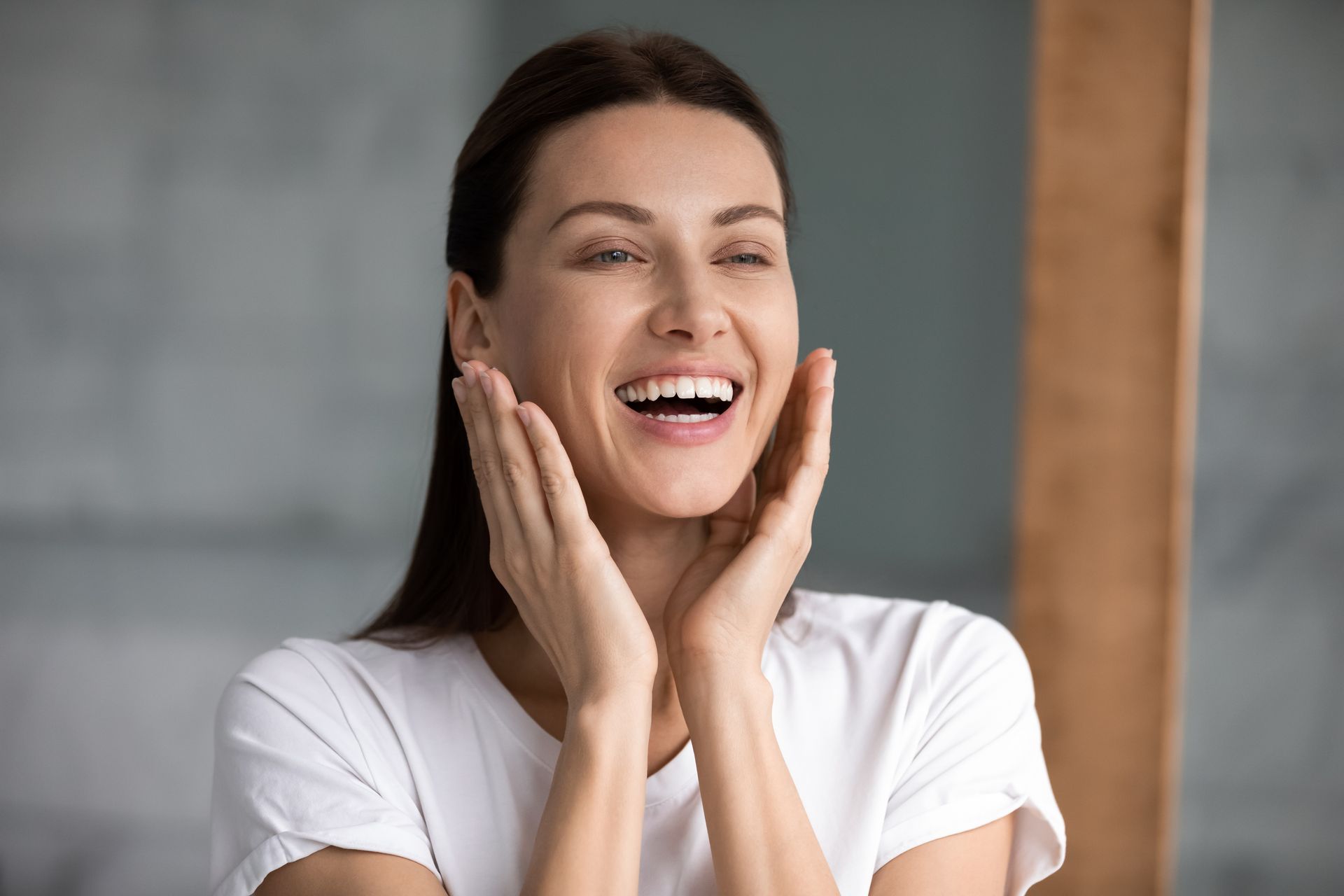 Woman in white t-shirt laughing, hands on cheeks, standing in front of a blurred background.