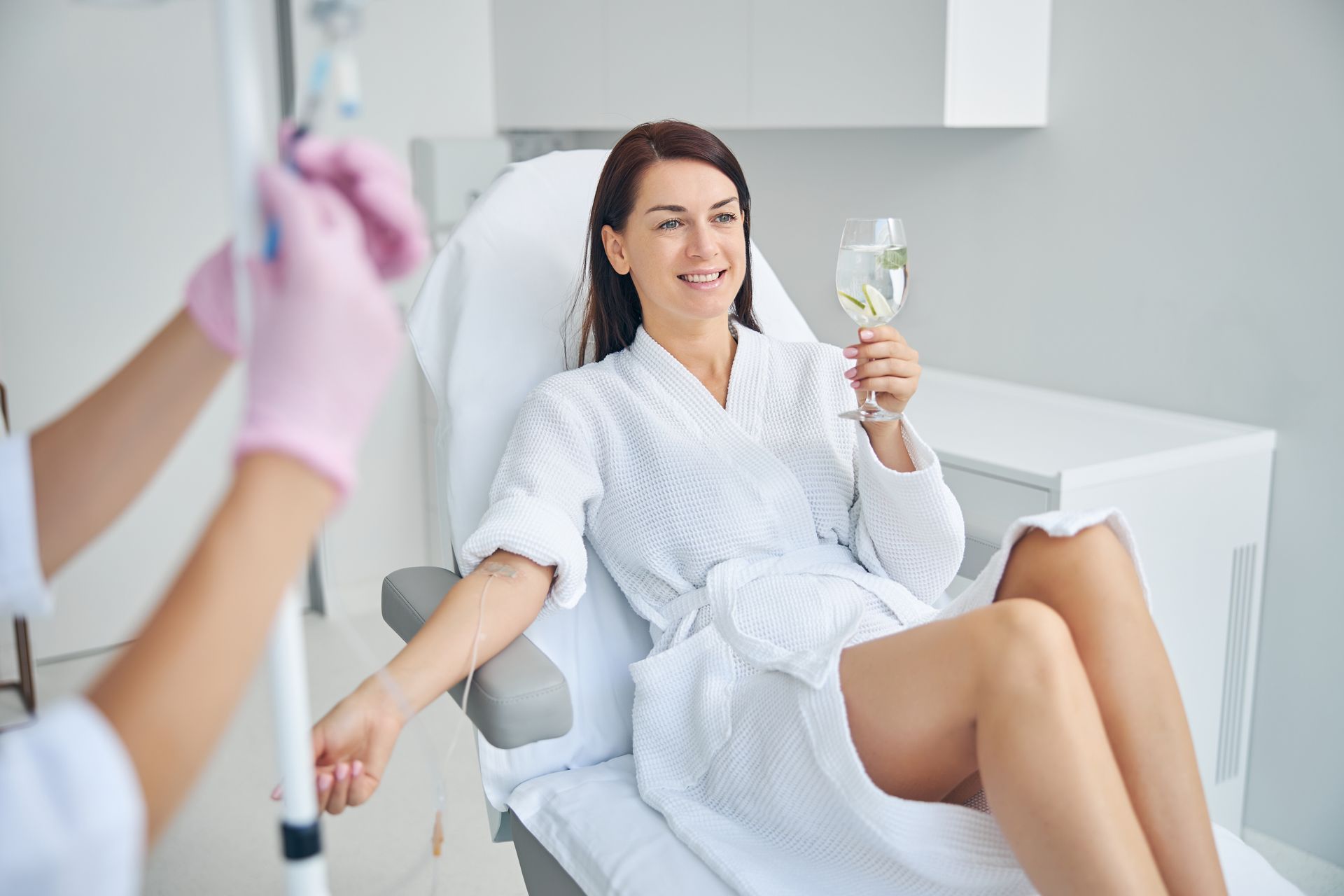Woman in robe receives IV drip, smiles, holds drink in spa-like setting.