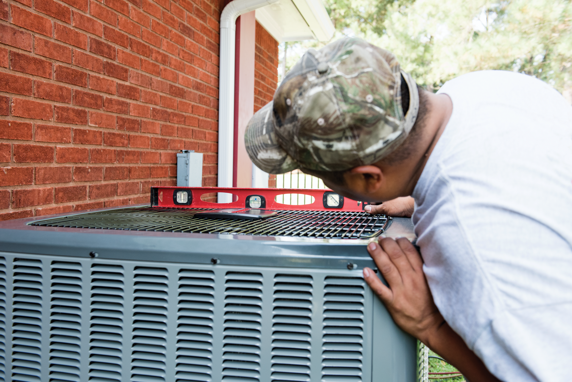 Man in camouflage cap inspecting an outdoor HVAC unit beside a brick house