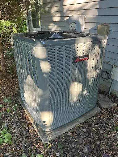 Outdoor HVAC condenser unit beside a house, with dappled sunlight and fallen leaves on gravel.
