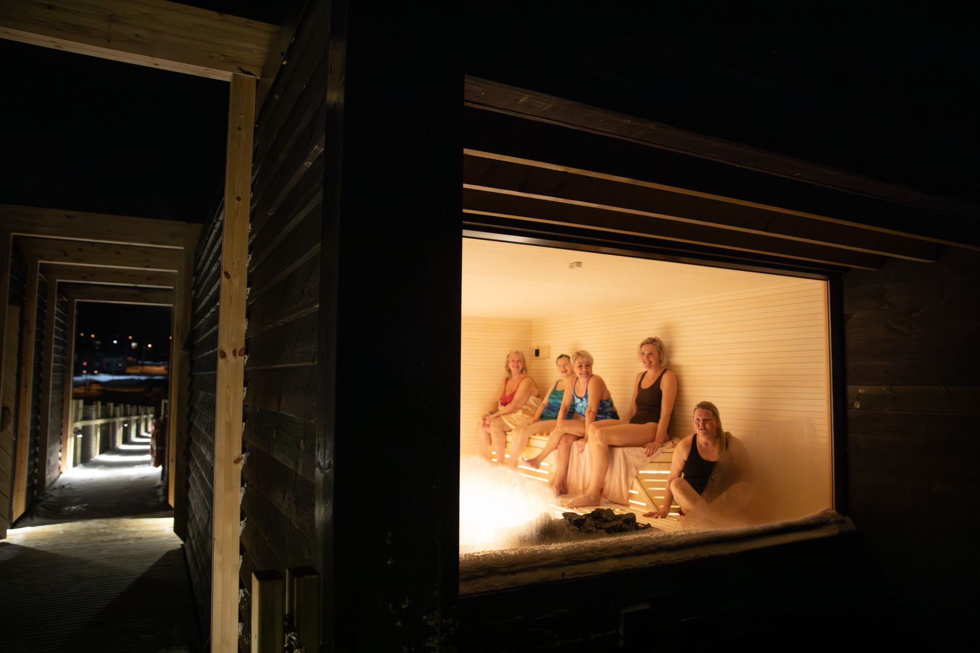 En kvinne titter ut av vinduet på en sauna i solnedgangen, hun smiler bredt og holder en badstulime av bjørk.