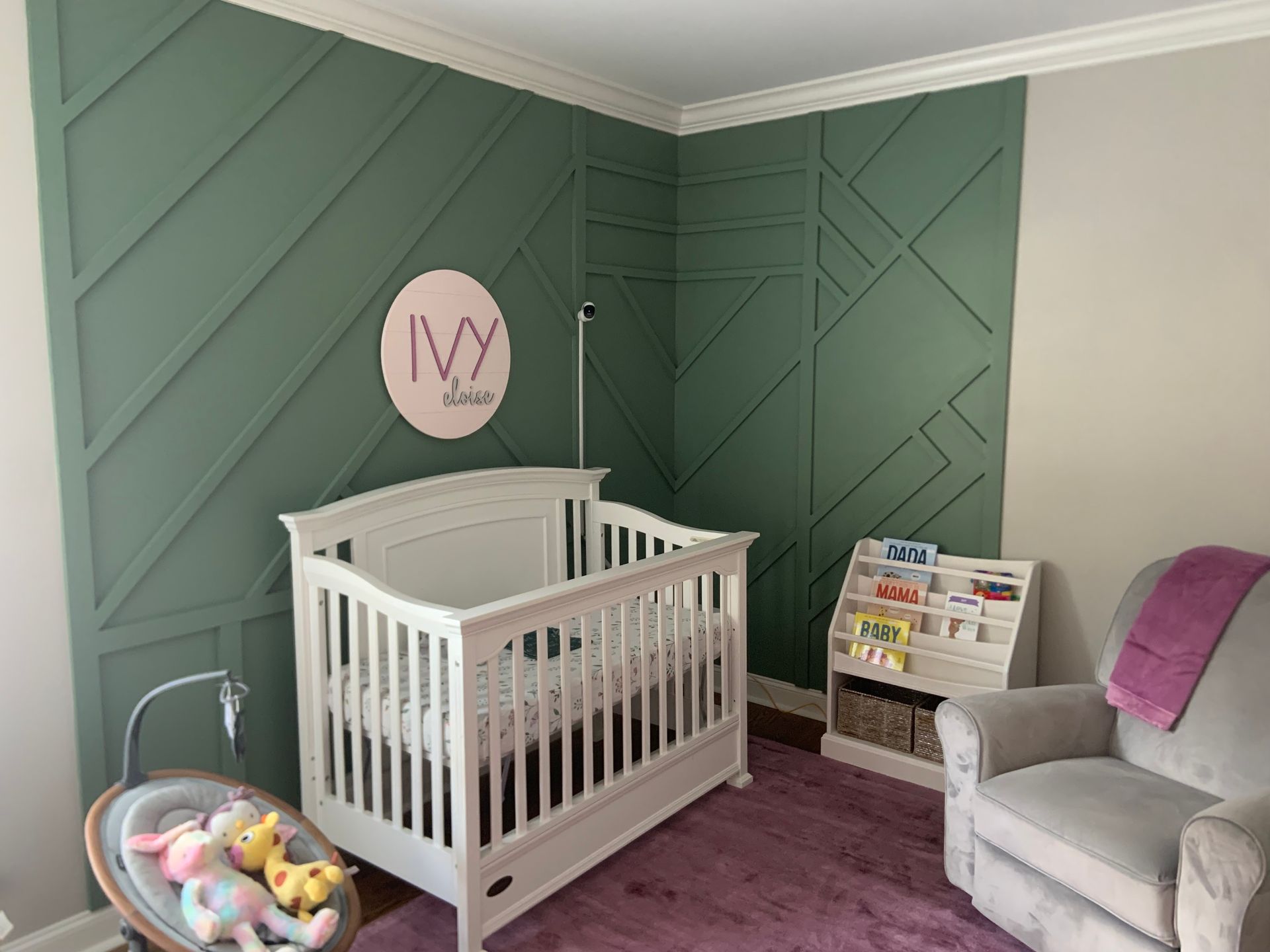 Nursery with white crib, green paneled walls, mauve rug, rocking chair, and toy basket.