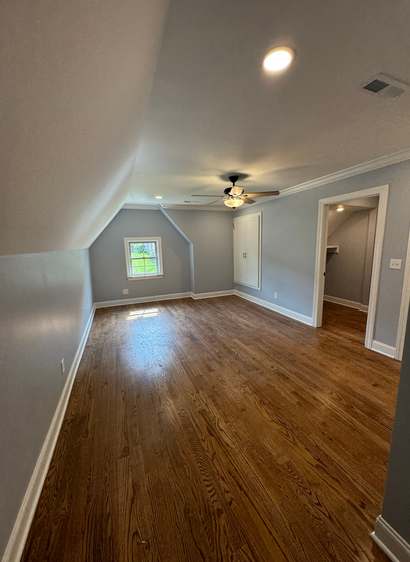 Empty gray attic room with wood floors, sloped ceiling, window, and ceiling fan.