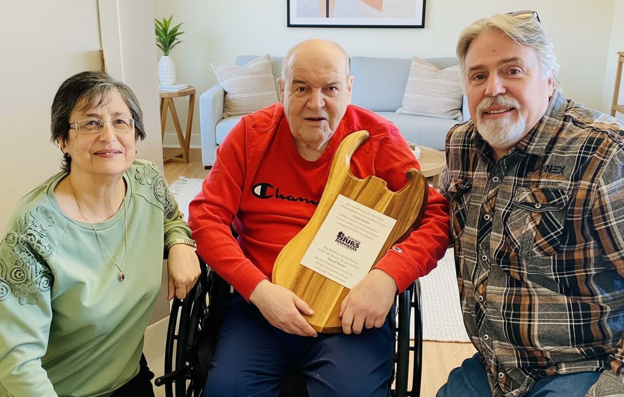 Une femme agenouillée à côté d’un homme en fauteuil roulant tenant une plaque en forme de guitare, avec un autre homme à sa droite.