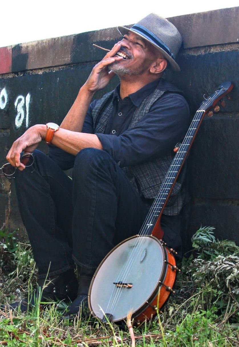 Photo may contain cigarillo, sunglasses, banjo, smiling man, watch, hat, cinder block wall