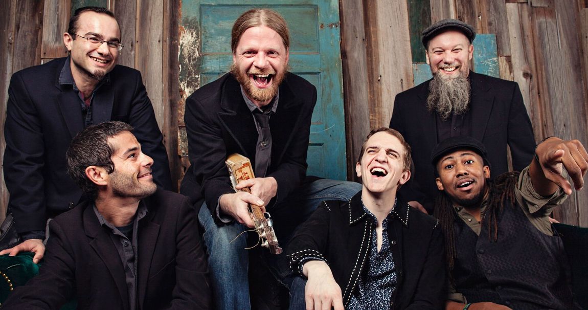 Photo contains laughing men posing with a cigar box guitar and sitting on a couch
