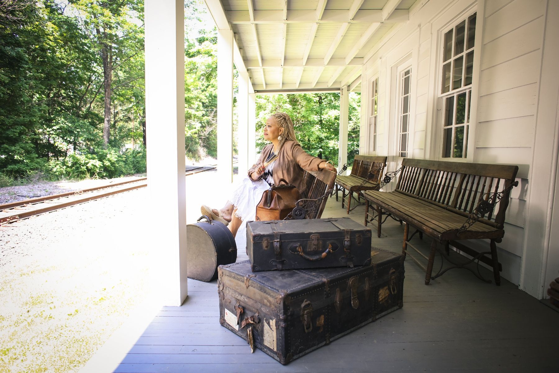 An elegant woman with long, blond dreadlocks, sitting outside a train station with suitcases next to her.