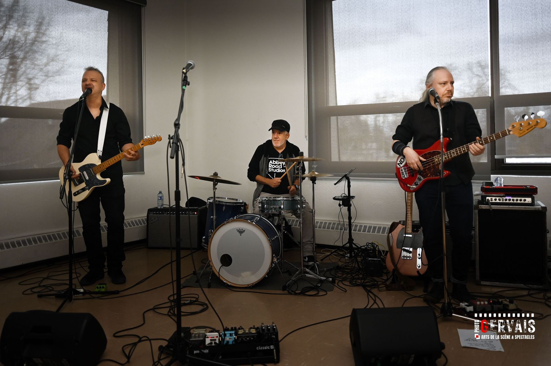 Drummer, guitar player and bass player playing in front of a window with blinds drawn